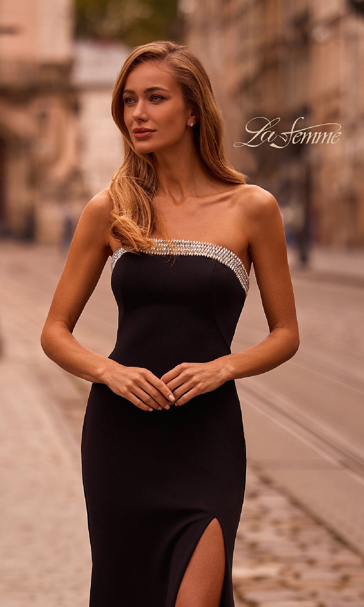 A woman stands on a cobblestone street, wearing the La Femme 32891 bead-trimmed long prom dress featuring a strapless neckline and thigh-high slit, with blurred buildings in the background.